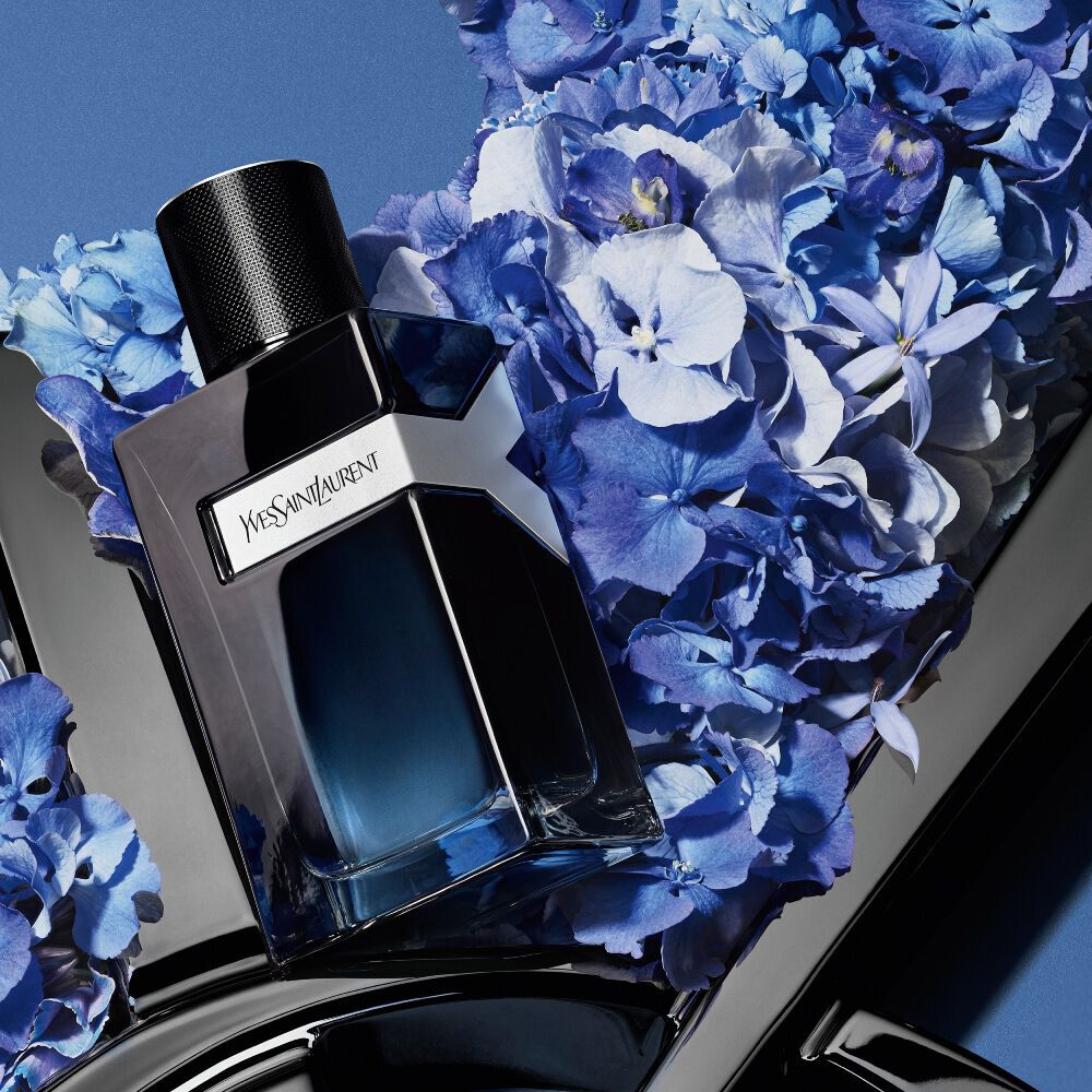 YSL Y Eau de Parfum fles op een glanzend zwart YSL-logo, omringd door levendige blauwe hortensia's tegen een diepblauwe achtergrond.