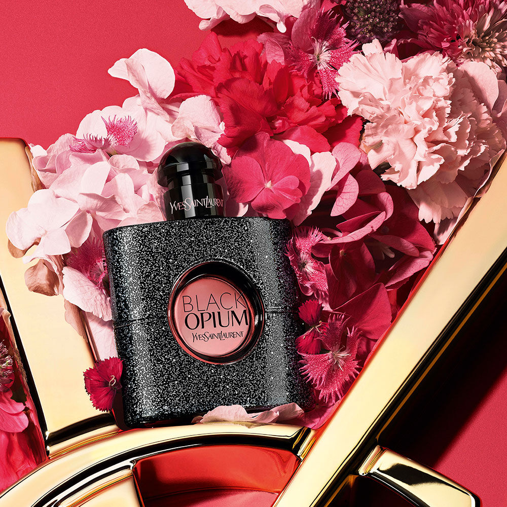 Black Opium Yves Saint Laurent glinsterde zwarte parfumfles omringd door roze en rode bloemen, gestyled op een rode achtergrond met een gouden YSL-logodetail.