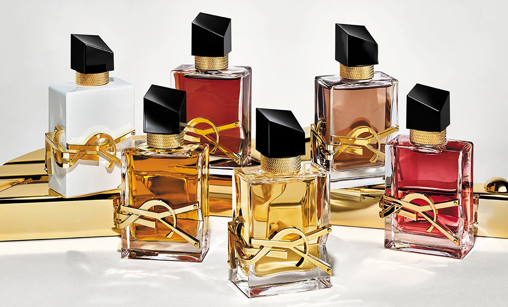 Six flacons de parfum YSL Libre sont présentés sur une surface dorée brillante. Les flacons présentent des variations de couleur du liquide et de finition : transparent, blanc, rougeâtre et brun. Tous arborent l'emblématique lettrage YSL doré et un bouchon noir incliné.
