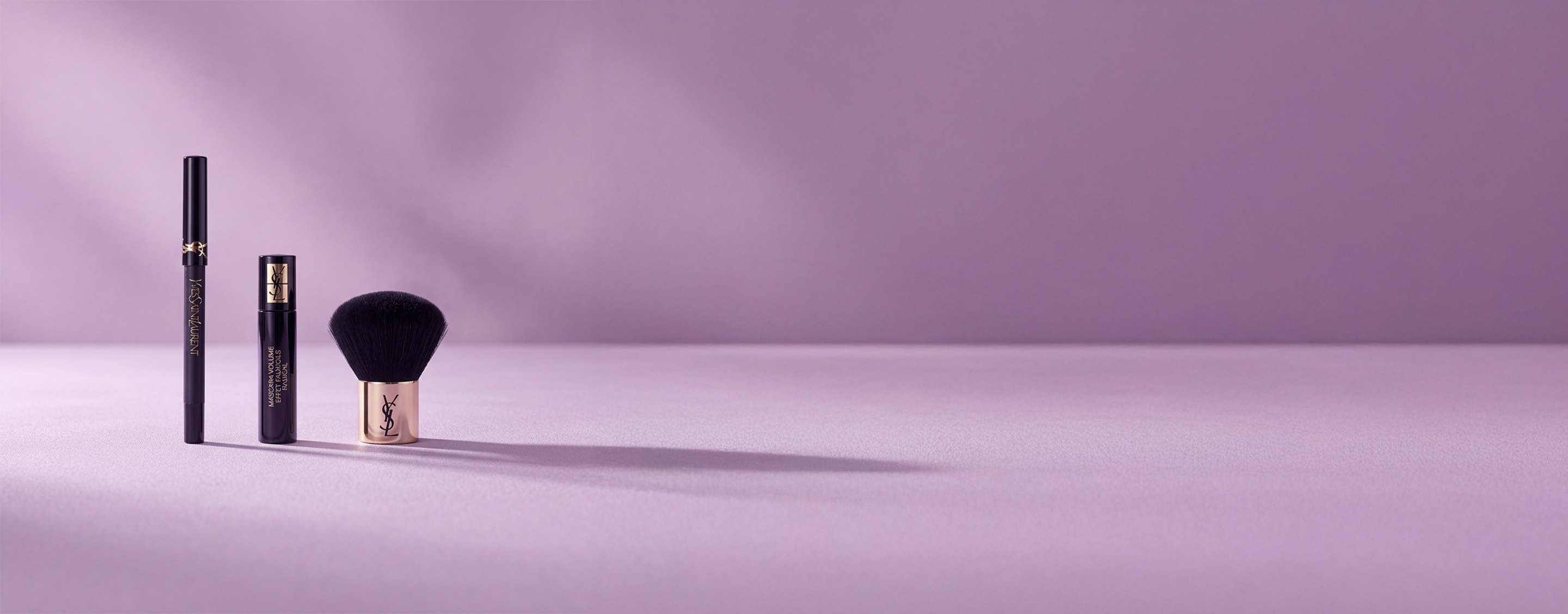 Trois produits de beauté Yves Saint Laurent, dont un mascara et un pinceau kabuki, sur un fond lavande doux.