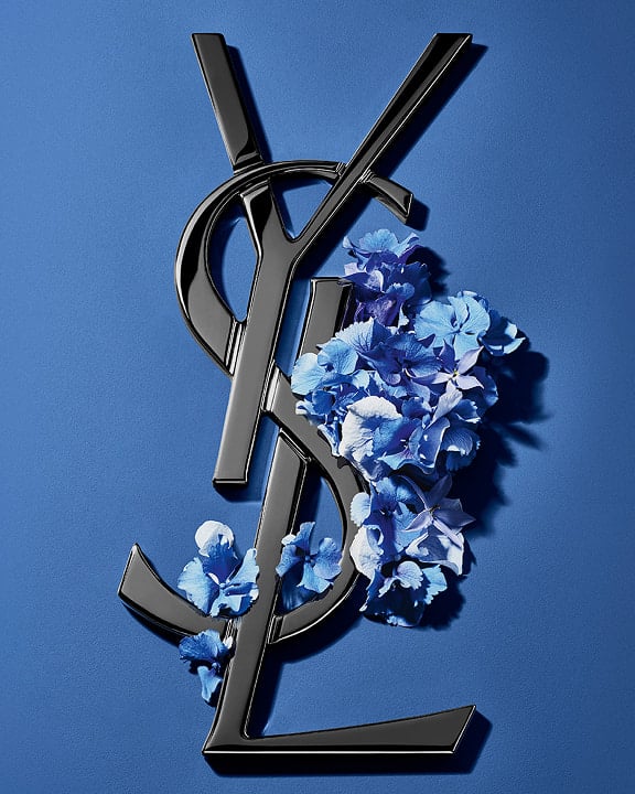 Zwart YSL Cassandre-logo versierd met levendige blauwe voorjaarsbloemen, tegen een serene blauwe achtergrond.