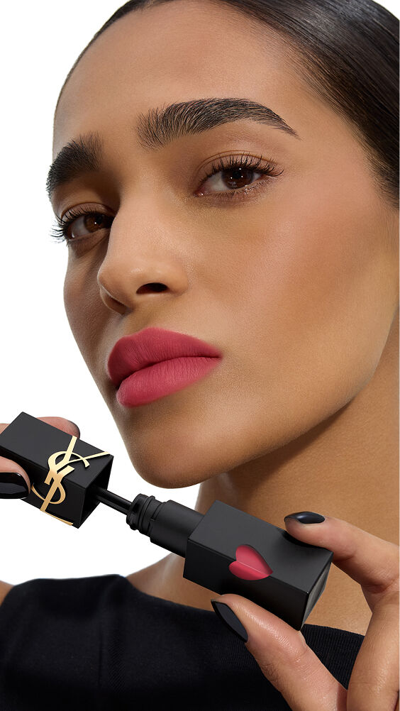 Modèle à la peau mate, lèvres d’un rose mat vibrant, tenant le flacon noir YSL THE INKS BLUR orné de son cœur rose.