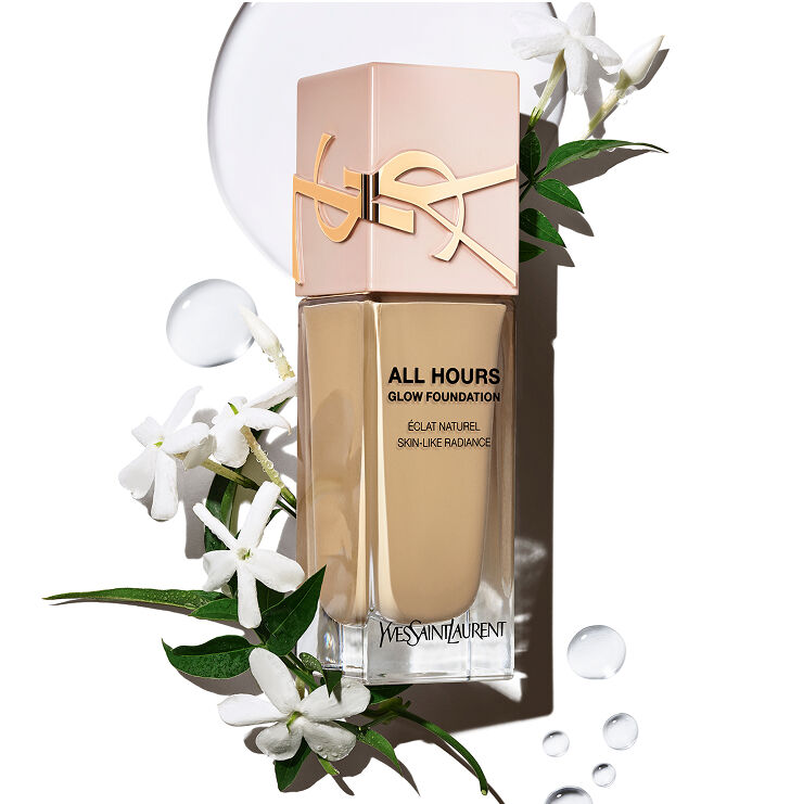 Bouteille de fond de teint YSL All Hours Glow avec 'Éclat Naturel Skin-Like Radiance' entourée de fleurs de jasmin blanches et de gouttelettes d'eau claire, suggérant des ingrédients naturels et une hydratation.