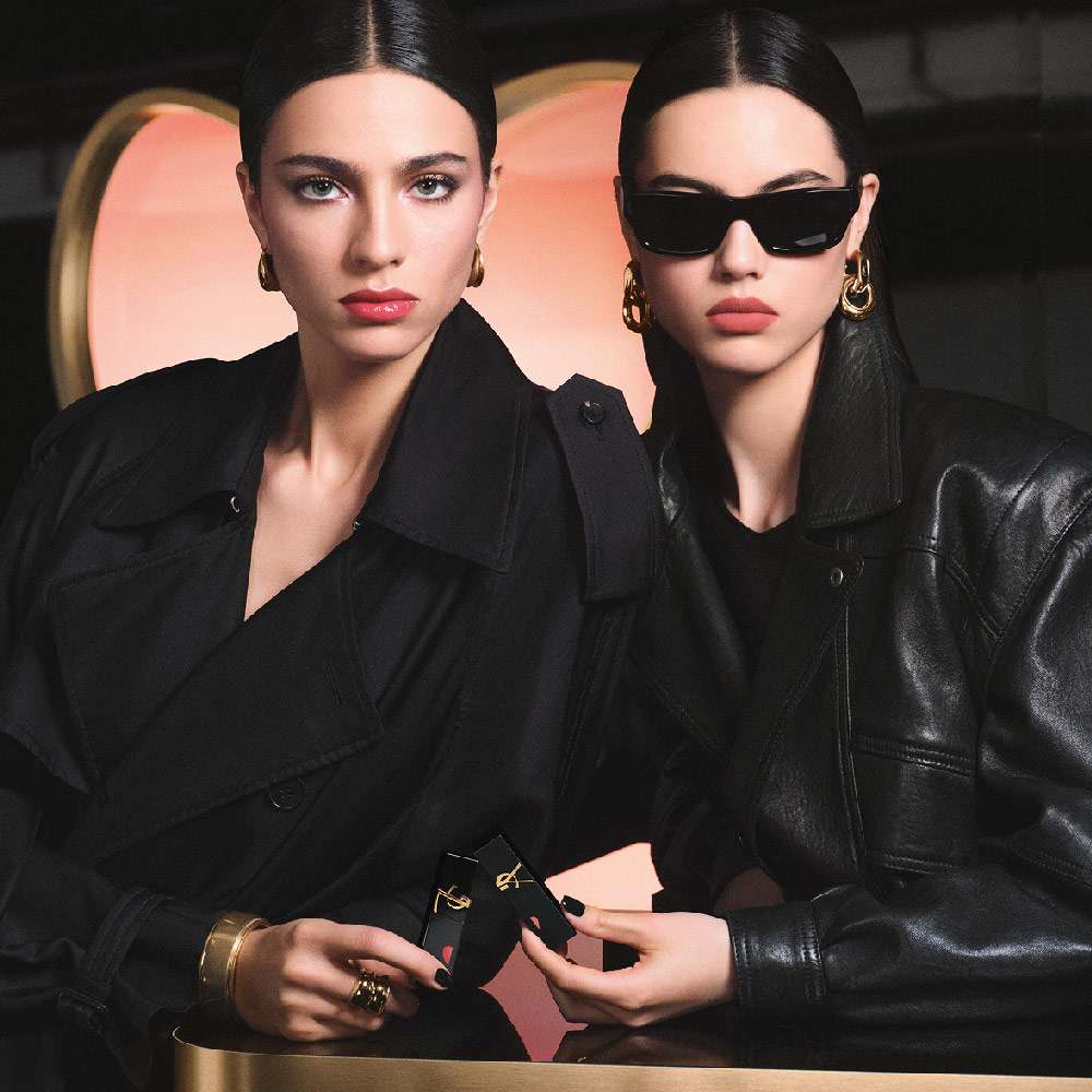 Deux mannequins chic en trench noir et veste de cuir tenant des rouges à lèvres YSL devant un immense décor en forme de cœur rose, affichant un look lèvres rouge brillant et un look lèvres rose mat velouté.