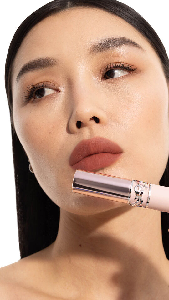 Model Mei J wearing YSL LOVENUDE Lip Blusher in Shade 3, holding the product near her rosy nude lips.