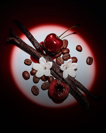 Gros plan sur des cerises rouges juteuses, des fleurs de jasmin, des gousses de vanille et des grains de café, sublimés par un éclairage dramatique pour Black Opium Eau de Parfum Over Red.