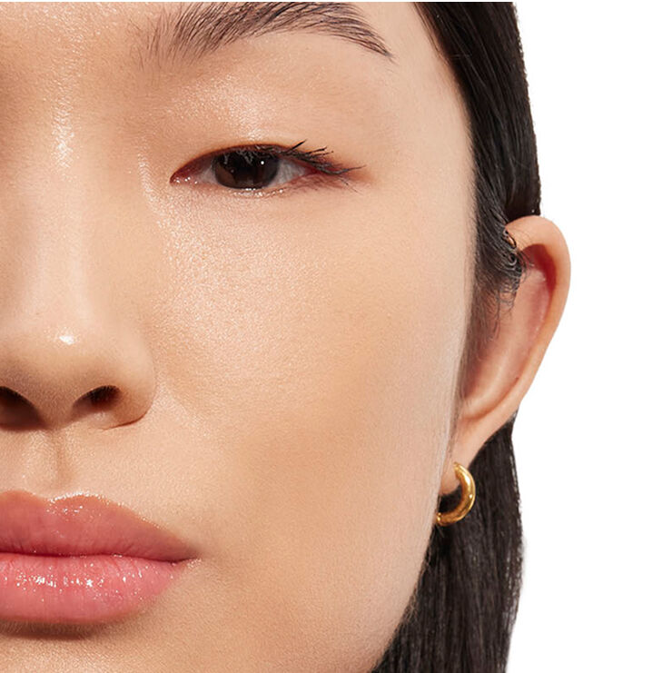 Gros plan détaillé du visage d'une personne, se concentrant sur la zone des yeux et des joues avec un maquillage d'apparence naturelle et une boucle d'oreille créole dorée, démontrant la luminosité de la peau.