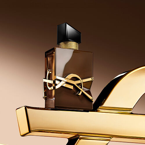 Flacon Libre Santal Couture brun glacé reflétant la lumière, posé sur une grande Cassandre YSL dorée.