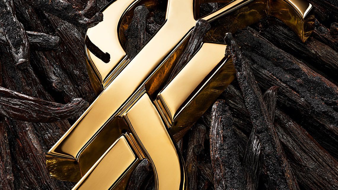Un logo YSL doré repose sur un lit de gousses de vanille foncées et texturées, ainsi que de fleurs d'oranger blanches, le tout rehaussé de paillettes d'or.