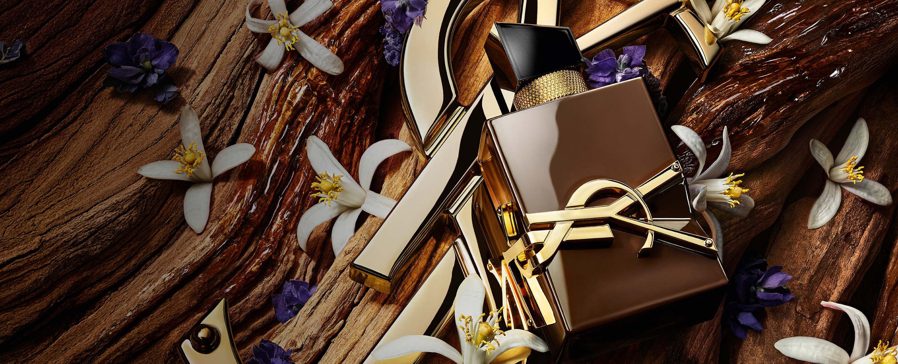 Flacon Libre Santal Couture brun glacé reflétant la lumière, posé sur une grande Cassandre YSL dorée.