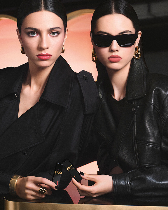 Deux mannequins chic en trench noir et veste de cuir tenant des rouges à lèvres YSL devant un immense décor en forme de cœur rose, affichant un look lèvres rouge brillant et un look lèvres rose mat velouté.