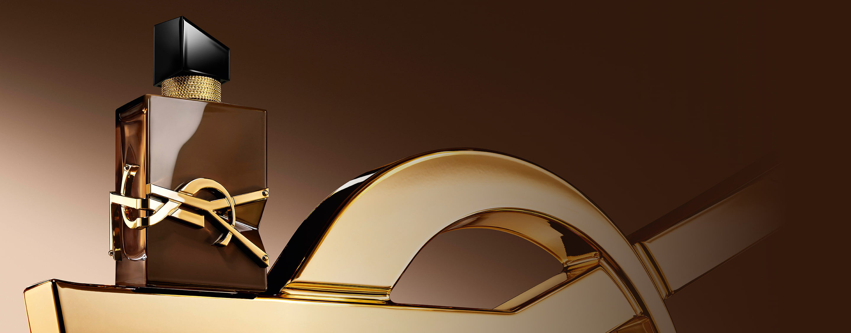 Flacon Libre Santal Couture brun glacé reflétant la lumière, posé sur une grande Cassandre YSL dorée.