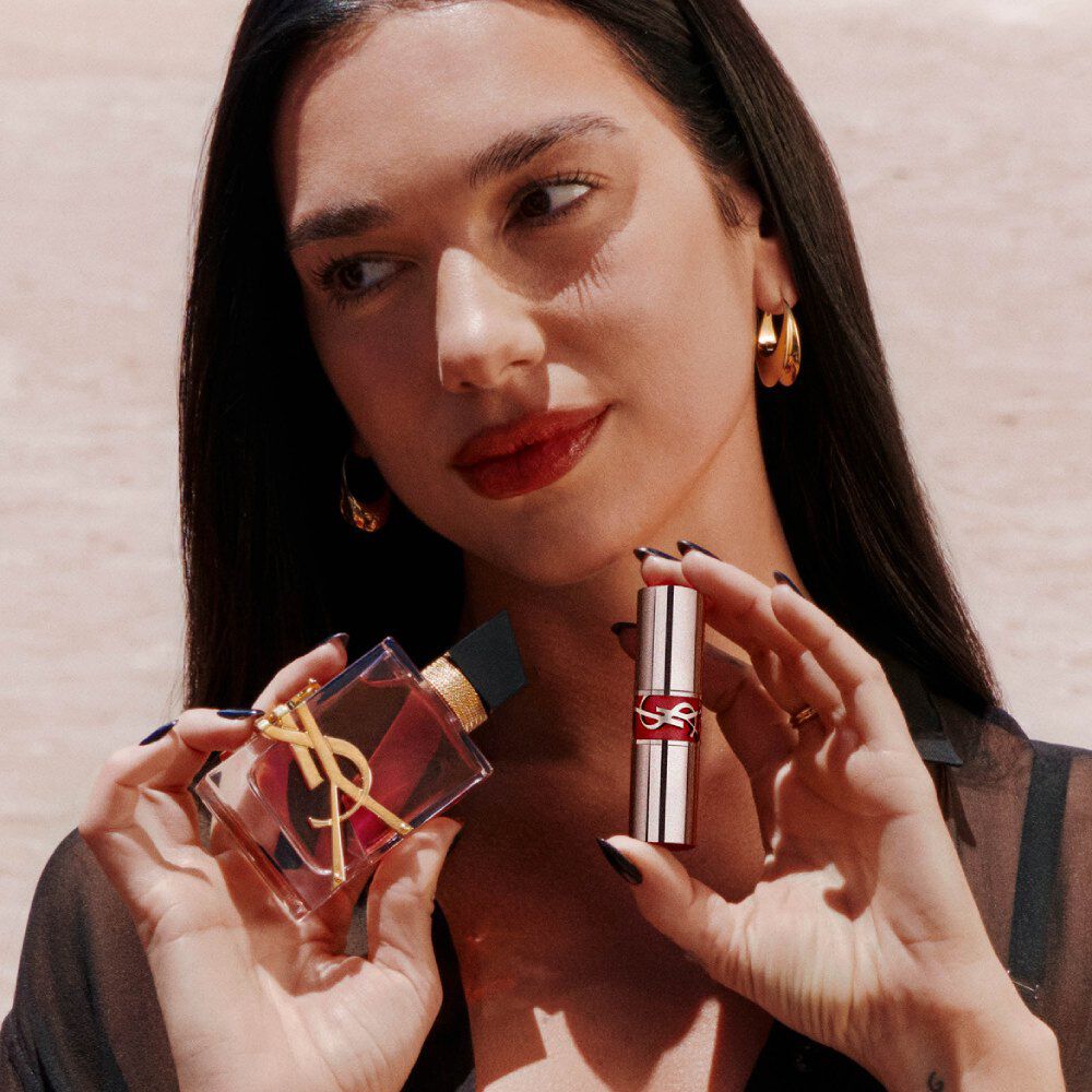 Dua Lipa tient Libre Eau de Parfum Berry Crush et un YSL Loveshine, avec des lèvres rouges et des boucles d'oreilles dorées.